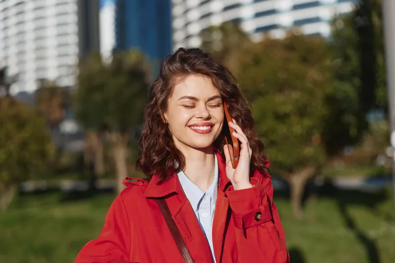 Young woman with curly brown hair smiles with closed eyes while talking on phone outdoors in a city park on a sunny day wearing a red coat and light blue shirt.