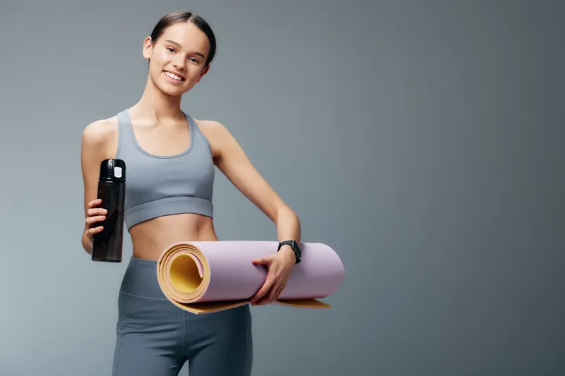 Fitness, woman, workout, training, yoga mat, bottle, exercise, gym, athletics, wellness. A fit woman in active wear holds a water bottle and rolled yoga mat, ready for routine training in a clean