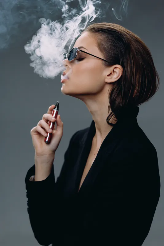 Young woman vaping with smoke in a dark studio, wearing sunglasses and black jacket, stylish and modern.