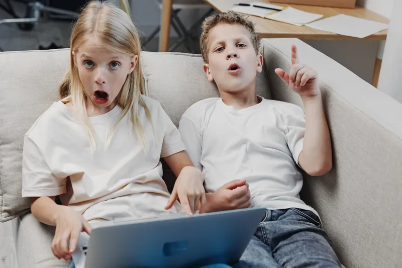 Two surprised children sit on a sofa using a laptop, reacting with shock and curiosity. Casual home setting with natural lighting shows genuine emotions and sibling interaction.