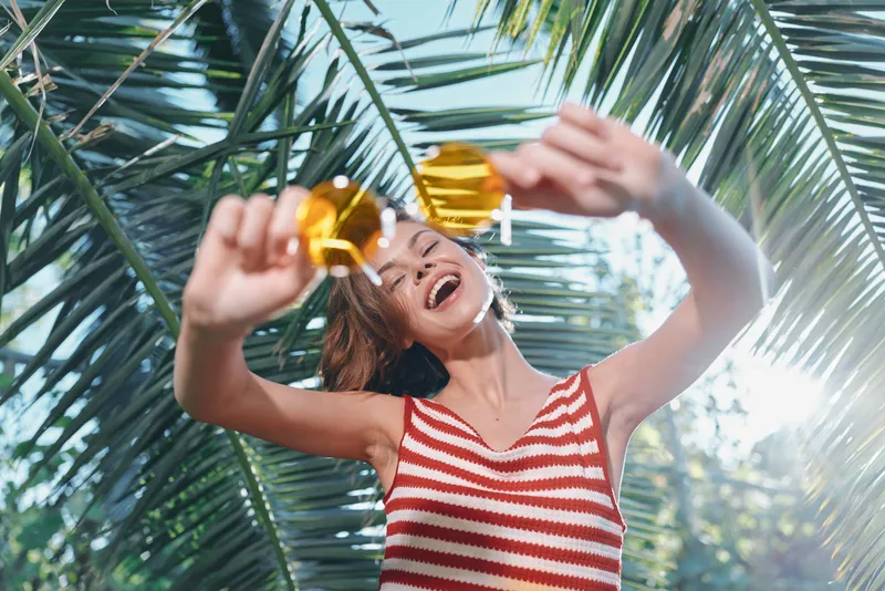 A joyful woman enjoys sunny tropical vibes as she lifts refreshing drinks beneath lush palm fronds, radiating energy, smiles, and carefree celebration in a vibrant outdoor scene.