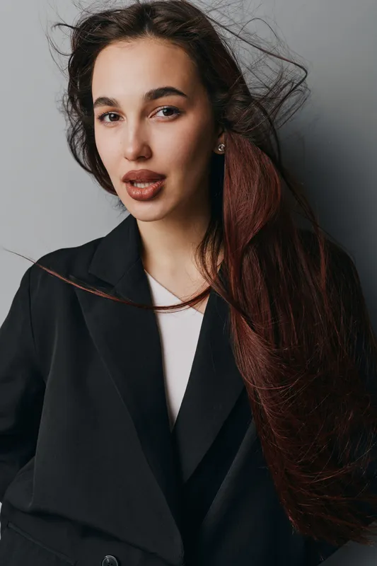 Young woman with long hair wearing black blazer and white shirt posing against gray background. Portrait of confident female model with natural makeup and loose hair in studio lighting.