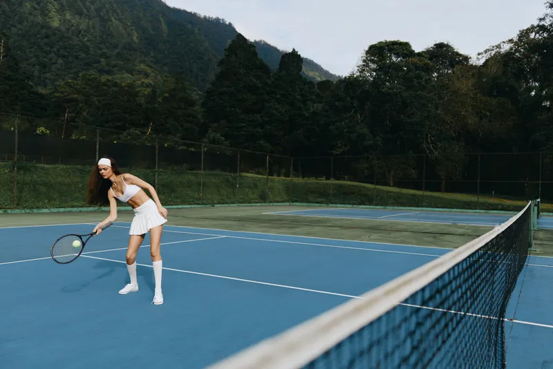 Young athletic woman in white tennis outfit playing on outdoor court, showing focus and determination, surrounded by lush greenery in summer