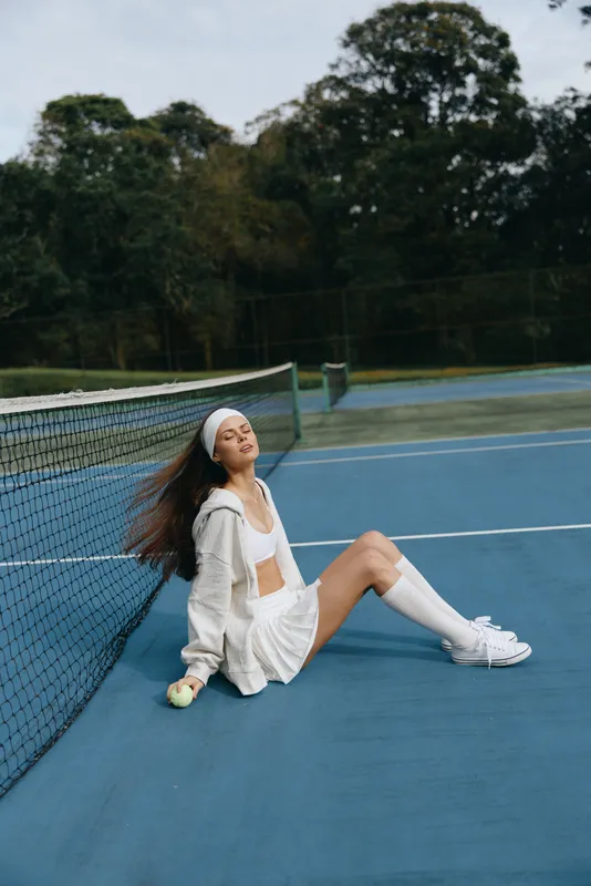 Young woman in white sportswear enjoying a moment of relaxation on a tennis court surrounded by greenery, conveying joy and serenity