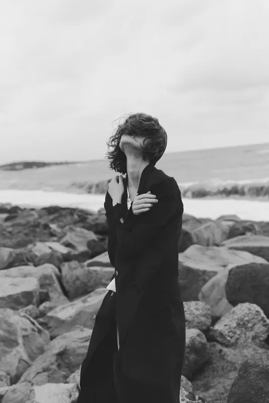 Wind swept woman in a long coat along a rocky shoreline, sea breeze, dramatic monochrome scene with movement and contrast.