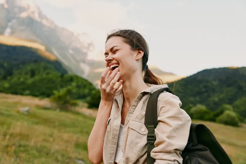 Woman lifestyle outdoors smiling in casual jacket with backpack on a hillside, mountains in the background. Concept for joyful travel, exploration, and everyday moments.