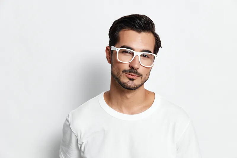 Handsome serious young man wearing white t-shirt and stylish glasses looking at camera with confident expression isolated on plain solid light background.
