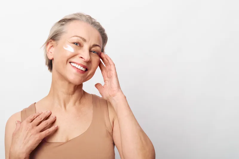 Happy mature woman with short hair applies face cream patches in bright studio. Concept of skincare, antiaging, selfcare, wellness, and natural beauty with glowing healthy skin.