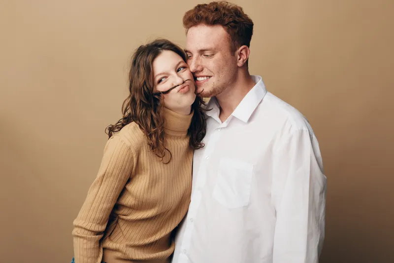 Young couple in casual clothes share a tender moment, smiling and embracing against a beige background. Concept of love, affection, and relationship happiness.