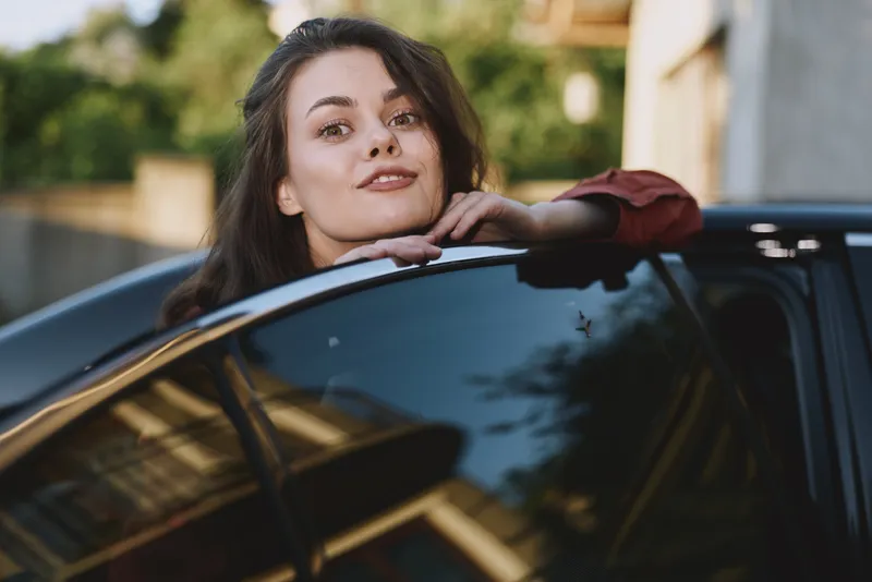 A confident young woman leans on a sleek car window, smiling softly in a sunlit urban setting, suggesting casual independence and everyday lifestyle