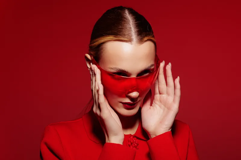 Stylish woman in red outfit and red sunglasses poses against red background. Fashion portrait with bold color contrast and modern accessories for trendy look.