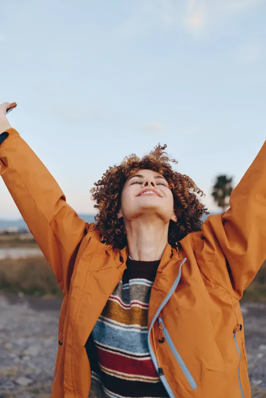 Young woman with curly hair, wearing an orange jacket, stands outdoors under a blue sky, arms raised, smiling, relaxed lifestyle, soft daylight, natural landscape