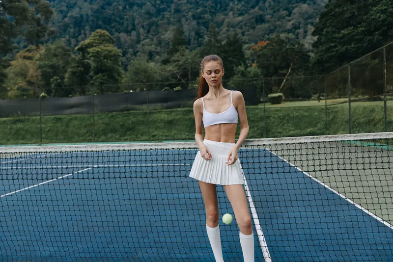 Young athletic woman in a white tennis outfit with a focused expression, standing on a court ready to serve, showcasing a competitive spirit and vibrant nature