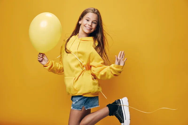 Girl in yellow hoodie and denim shorts holding yellow balloon jumping with smile against yellow background. Youth, happiness, casual, lifestyle, joy and party concept.