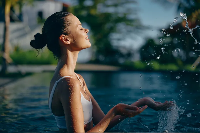 Lifestyle portrait of a woman relaxing by a sunlit pool, feeling the spray from a fountain, enjoying warm breeze, serene moment, casual summer scene, outdoor leisure by water, candid mood.