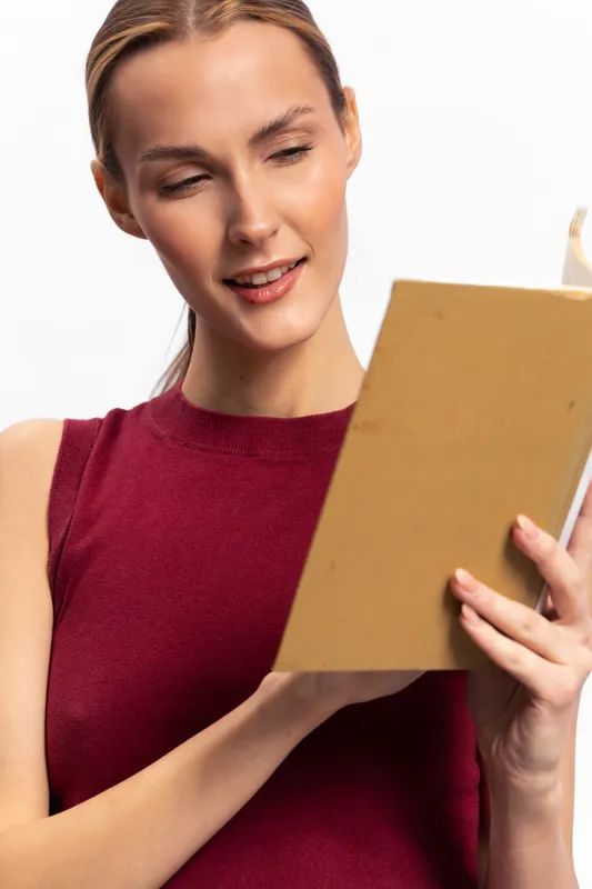 Young woman in a red sleeveless top reading a book with a gentle smile. Isolated on white background, concept of leisure, education, relaxation, and knowledge.
