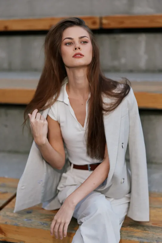 Fashion woman posing confidently in beige suit jacket and white top with long brown hair sitting on wooden bleachers outdoor