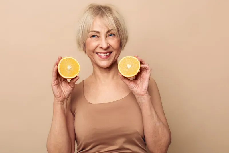 Smiling mature woman with short blonde hair holds two halves of fresh orange near face. Beige background, natural light, concept of healthy lifestyle, vitamin C, wellness, and positive energy.