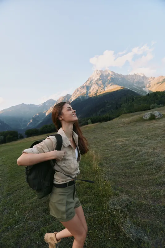 Cheerful woman hiking outdoors, backpacker smiling up at the mountains, sunlit meadow, casual attire, sense of adventure, nature colors, expansive landscape