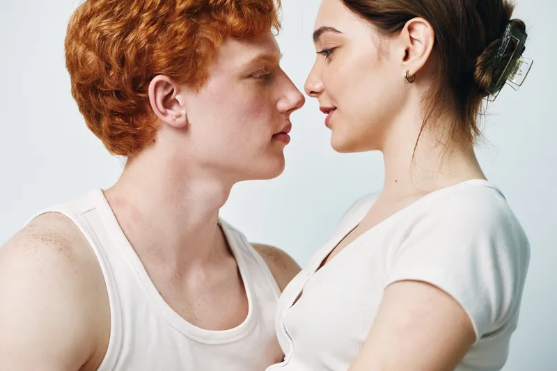 Couple, young man and woman in close intimate pose facing each other with gentle expressions in white clothing against light background.
