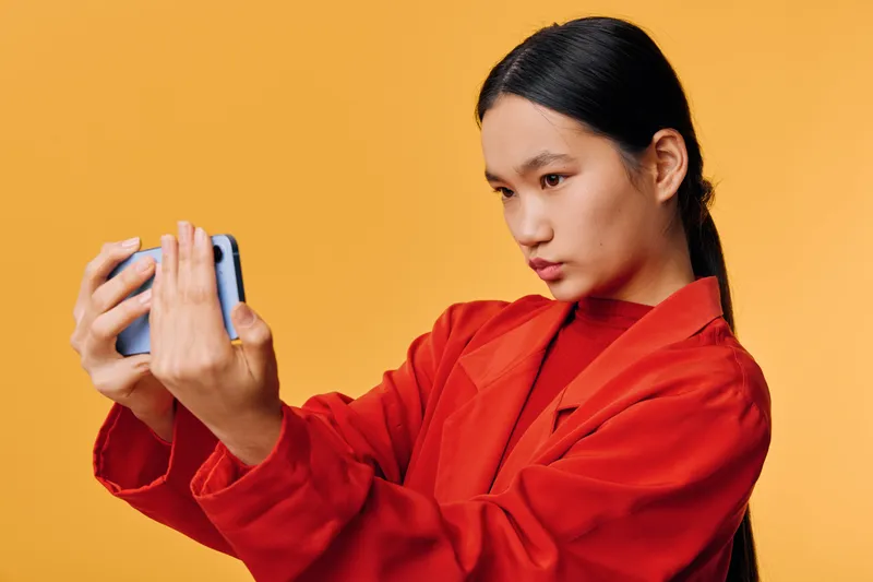 Young woman with long black hair wearing red jacket is taking a selfie with smartphone on yellow background, focused face and casual pose for social media or personal photo.