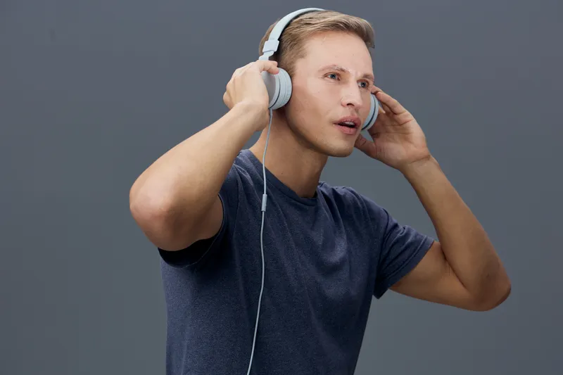 Young man with short blond hair, wearing a dark blue t-shirt, listening to music on large white headphones, looking thoughtfully to the side, isolated on plain gray background studio portrait. People 