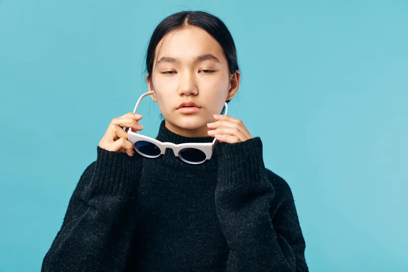 Young woman with closed eyes holding white sunglasses in hand wearing black sweater standing against blue background. Female model with minimal makeup and calm expression in studio portrait.
