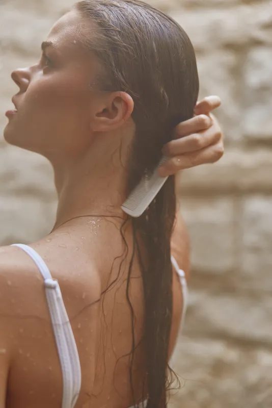 young woman combing wet hair in shower with white strap top, natural skin, soft light and blurred stone wall background, intimate morning self-care moment