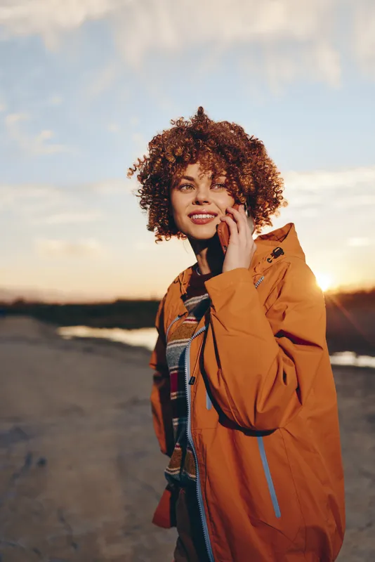 Woman outdoors, lifestyle portrait, curly hair, smiling in sunset light, wearing orange jacket, standing on road, relaxed pose, warm evening sky, natural colors