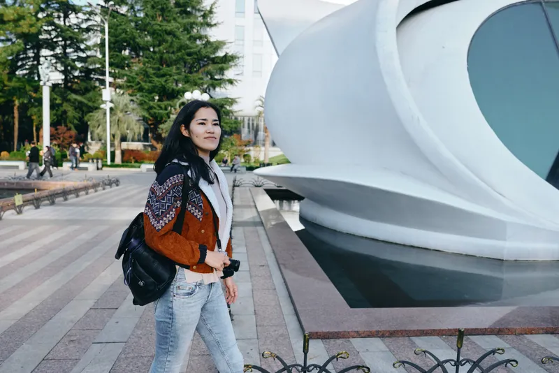 Woman with camera strolls beside a modern architectural sculpture in an outdoor urban setting, casual fashion, backpack, capturing a travel vibe and street photography mood