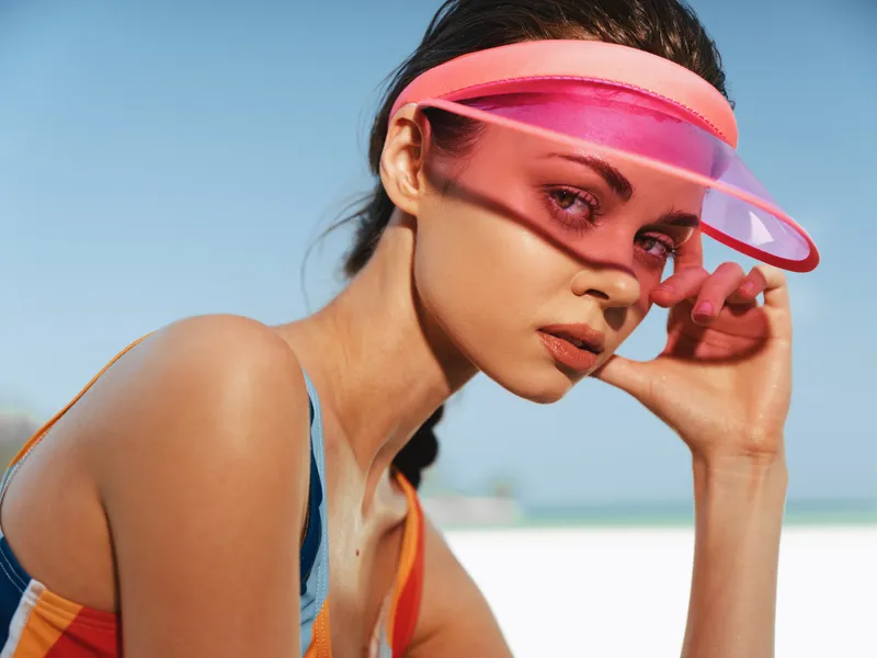 Close up portrait of a stylish woman on a sunny beach, wearing a pink translucent visor, vibrant colors, smooth skin, and a contemplative gaze. Lighting highlights texture and summer mood.
