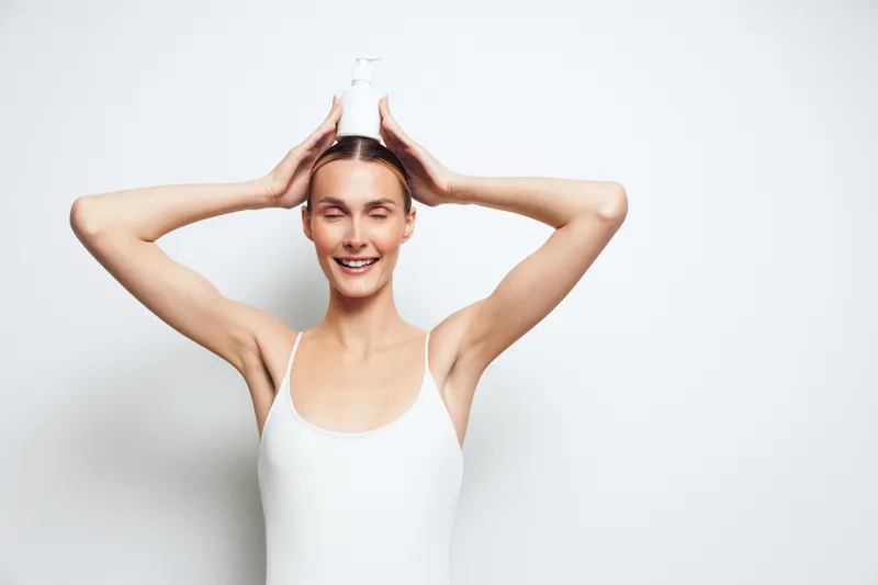 Smiling woman in white tank top holding a glass milk bottle on her head, standing against a white background. Concept of dairy, health, nutrition, and happiness with soft lighting.