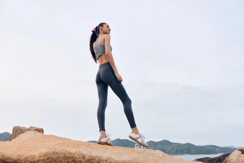 Young athletic woman in activewear smiling on rocky shore against calm ocean landscape, representing fitness, health, and outdoor lifestyle