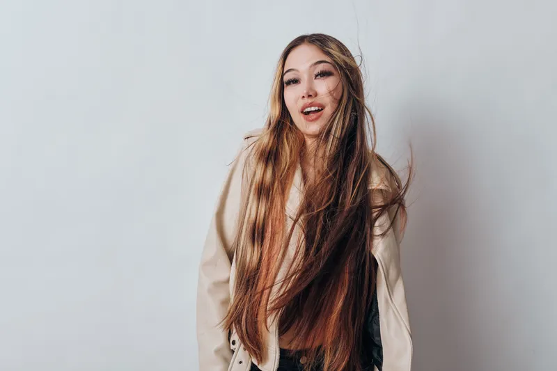 Young woman with long brown hair wearing a beige jacket posing against white background, smiling and looking at camera in casual style portrait photography.