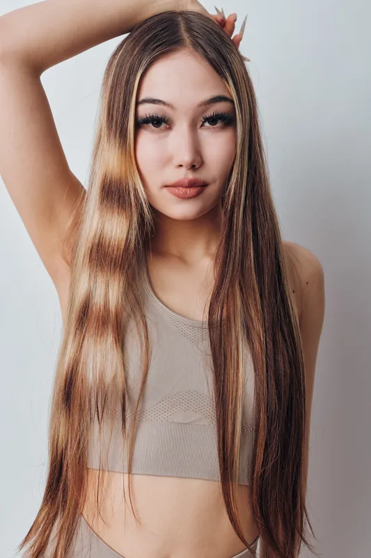 Young woman with long straight hair posing with hand behind head wearing beige top. Beauty, confidence and fashion style on light background for portrait and model.