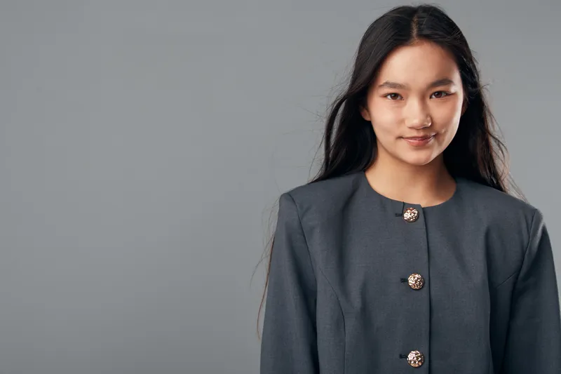 Portrait of confident young woman with long dark hair smiling in studio with plain gray background wearing elegant dark buttoned jacket casual closeup fashion style.