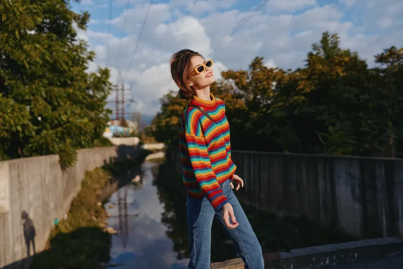 young woman early 20s, rainbow sweater, sunglasses, urban bridge, casual jeans, walking pose — smiling and relaxed in colorful knit on canal walkway, golden hour travel lifestyle portrait outdoors
