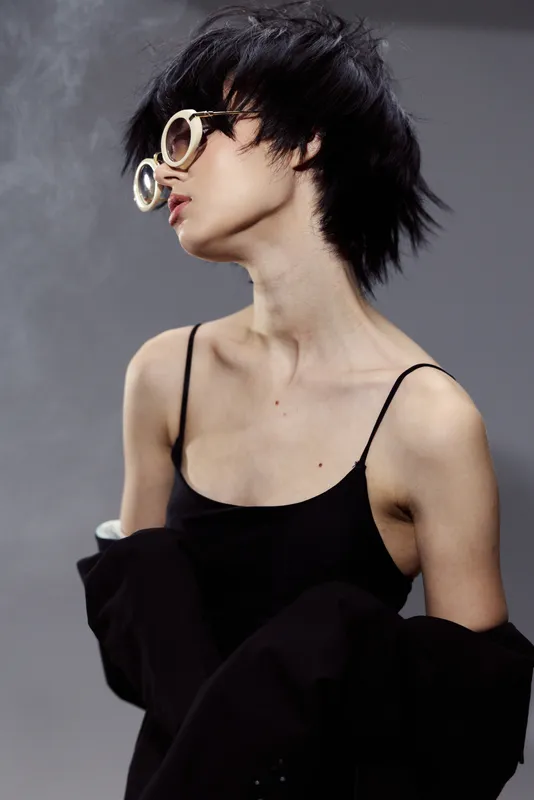 Stylish young woman with short hair wearing trendy round glasses and a black outfit against smoky dark background