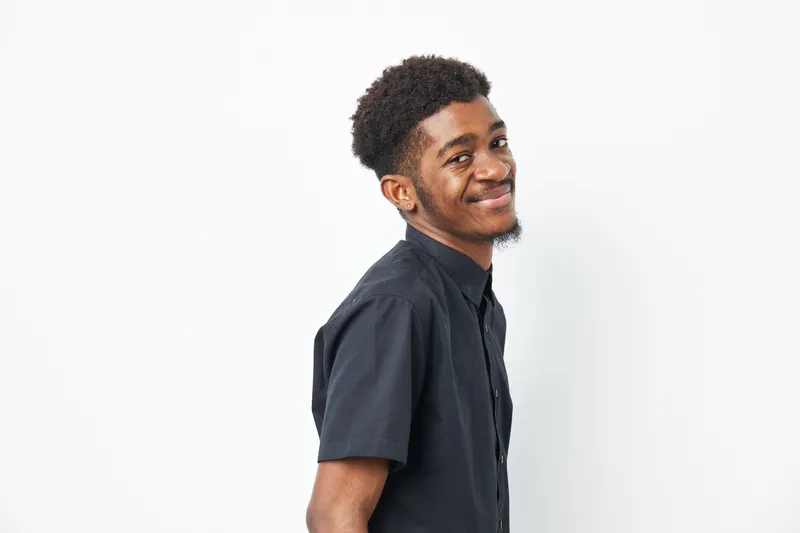 Portrait of a cheerful young African American man smiling, showcasing his stylish black shirt and confident expression. Bright, inviting atmosphere enhances the joyful mood.