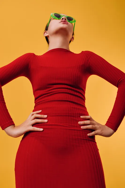 Woman in red long sleeve dress with hands on hips wearing green sunglasses posing confidently against vibrant yellow background. Fashion, style, attitude, modern and colorful portrait.