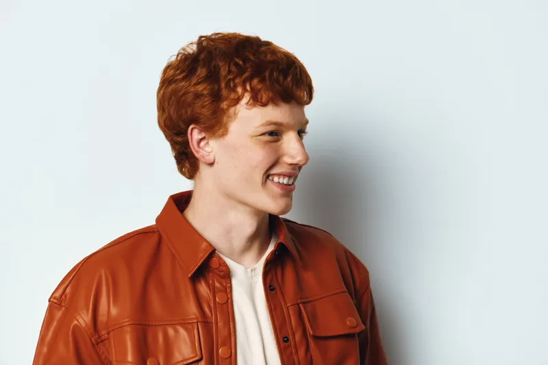 Young man with red hair and freckles wearing brown leather jacket smiling and looking sideways on white background. Casual portrait of male teenager with natural expression and hairstyle.