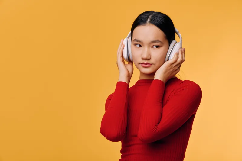 Young woman wearing headphones with closed eyes listening to music, standing in front of warm yellow background, emotional and peaceful moment with casual red outfit and dark hair.