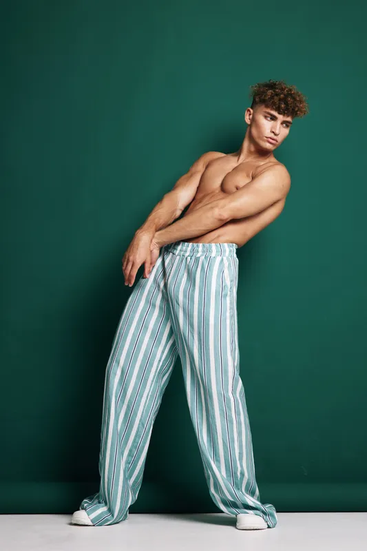 Young man with curly hair poses shirtless wearing loose striped pants and white sneakers against dark green studio background showing modern urban fashion and casual confidence.