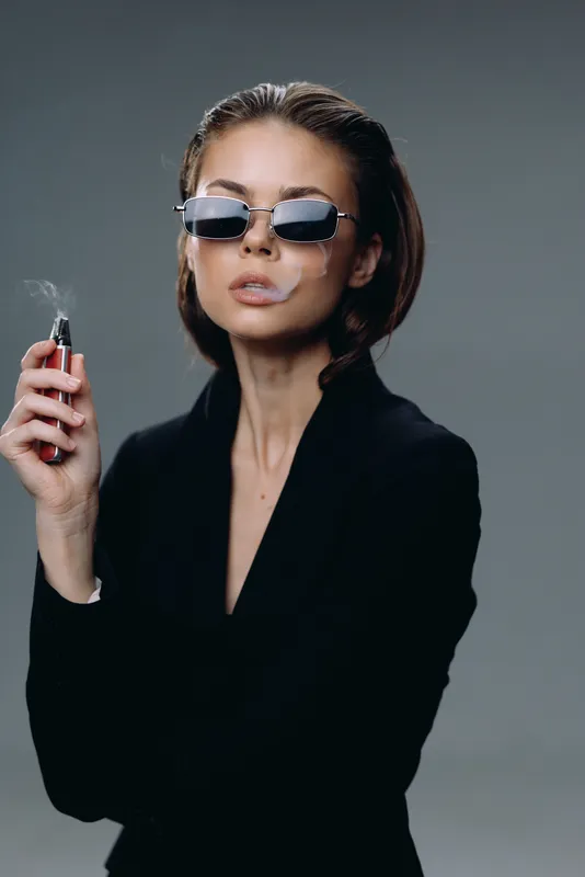 Stylish woman with short hair wearing sunglasses and black outfit smoking vape with smoke around her face