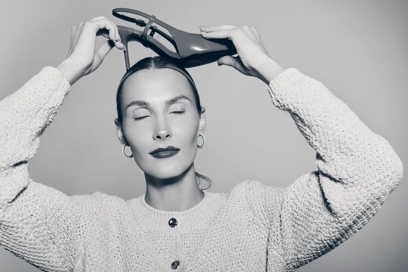 White woman with pale skin holds high heel shoe on head with closed eyes in studio. Concept for fashion, style and beauty.