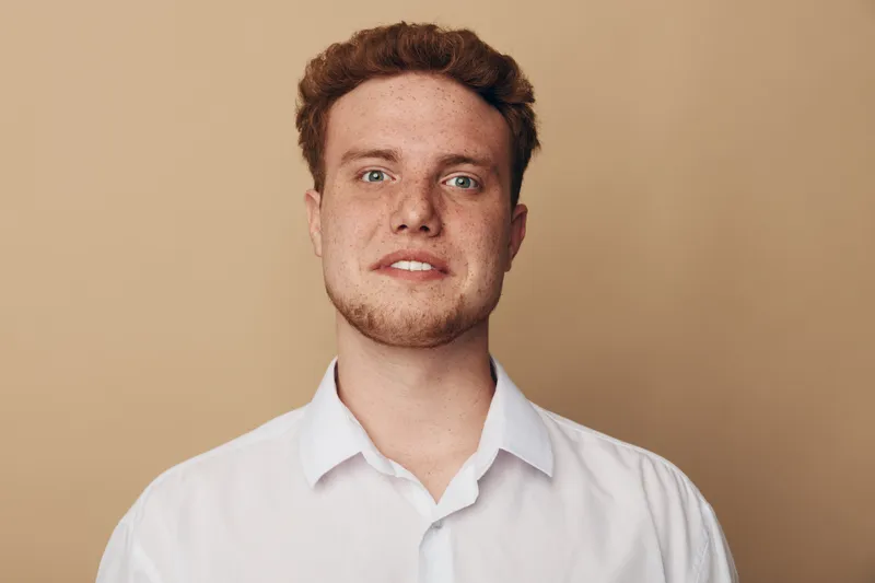 Young man with red hair and freckles wears white shirt, looking at camera with neutral expression on beige background. Concept of portrait, casual style, and natural appearance.