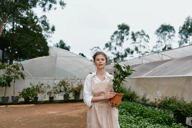 Young woman holding a potted plant in a greenhouse, wearing a light colored apron, exuding a serene expression, surrounded by greenery and soft natural light