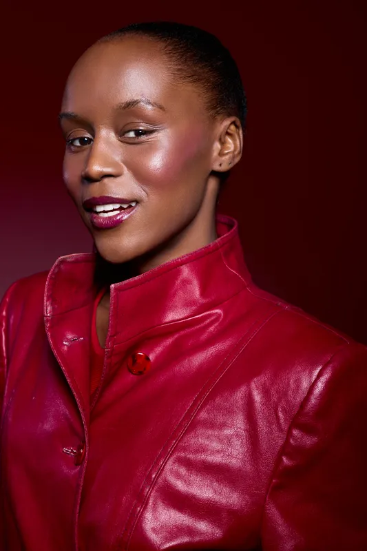 Confident African woman with dark skin wearing a red leather jacket smiles softly against a dark red background. Studio portrait highlighting fashion and elegance with warm lighting.