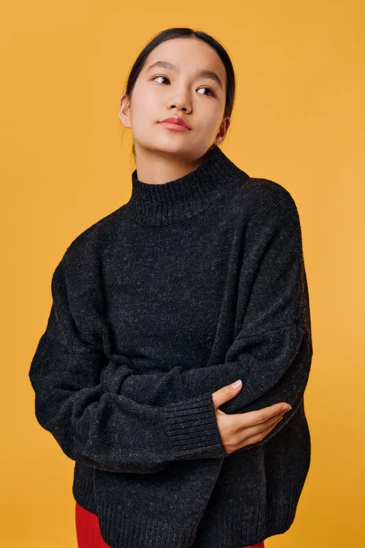 Young woman in black sweater poses with crossed arms against yellow background, looking to side with thoughtful expression. Casual portrait of female model in cozy knitted clothing studio shot.