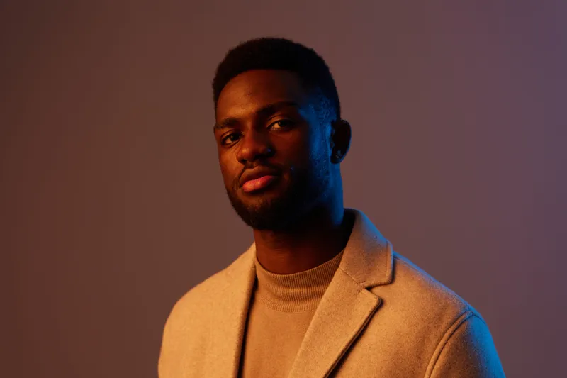 Portrait of a confident Black man in a stylish beige coat, showcasing a thoughtful expression against a gradient background, ideal for fashion and lifestyle imagery